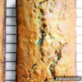 A golden brown loaf of gingerbread-zucchini bread rests on a cooling rack, its inviting aroma filling the air, with a charming crack adorning its top.