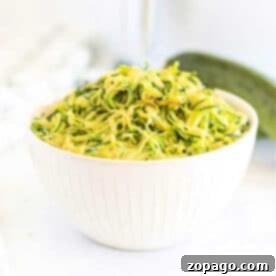 White bowl filled with shredded zucchini on a white surface, perfect for making air fryer zucchini noodles, with a fresh zucchini in the background.