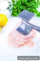 Raw chicken breast being tenderized with a metal mallet, surrounded by eggs, parsley, and a lemon. Wondering how long to bake chicken cutlets? This setup is perfect for creating the tender base you’ll need before popping them in the oven for delicious results.