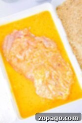 Raw chicken breast marinating in a yellow mixture, next to breadcrumbs in a white tray, waiting to become delicious baked cutlets. For perfectly cooked cutlets, consider how long to bake chicken cutlets at your desired temperature and enjoy a tender culinary delight.