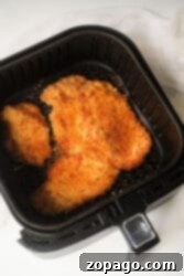Breaded chicken cutlets, reminiscent of air fryer chicken katsu, are cooking in an air fryer basket. The cutlets are golden brown and crispy, set against a black basket surface on a white background.