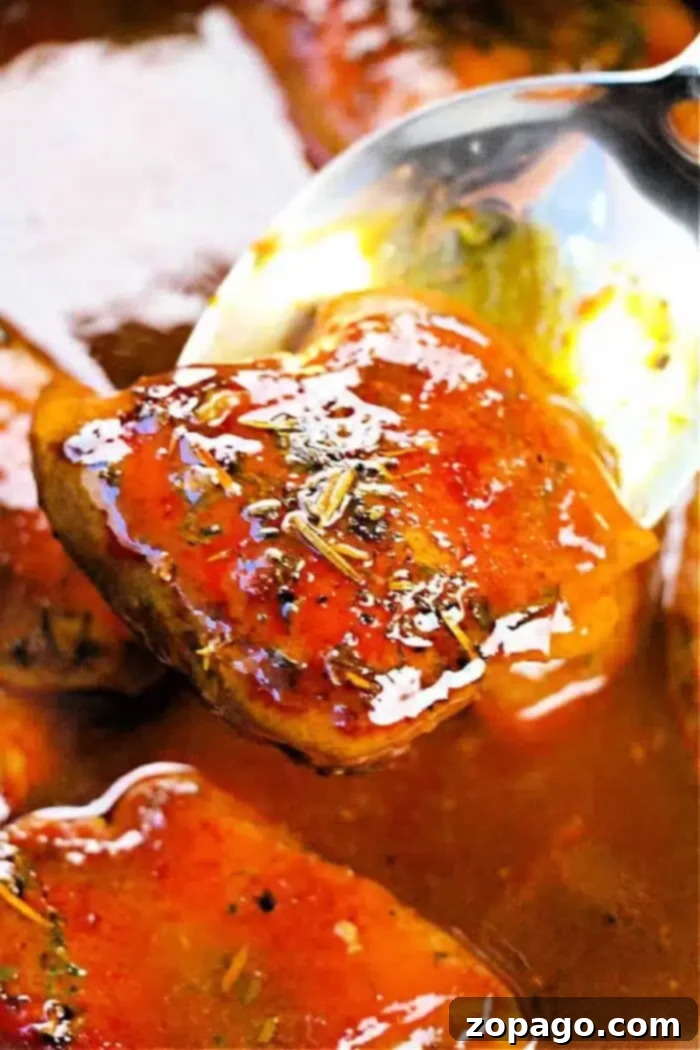 Effortless Slow Cooker Boneless Skinless Chicken Thighs 8 A close-up of a spoon lifting a piece of boneless, skinless chicken thigh coated in a glossy, reddish-brown sauce. The sauce appears thick and well-seasoned with visible herbs. More tender pieces simmer in the crockpot’s sauce in the background.