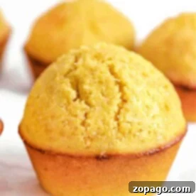 Close-up of a golden-brown cornmeal muffin with a slightly cracked top, sitting on a white surface.