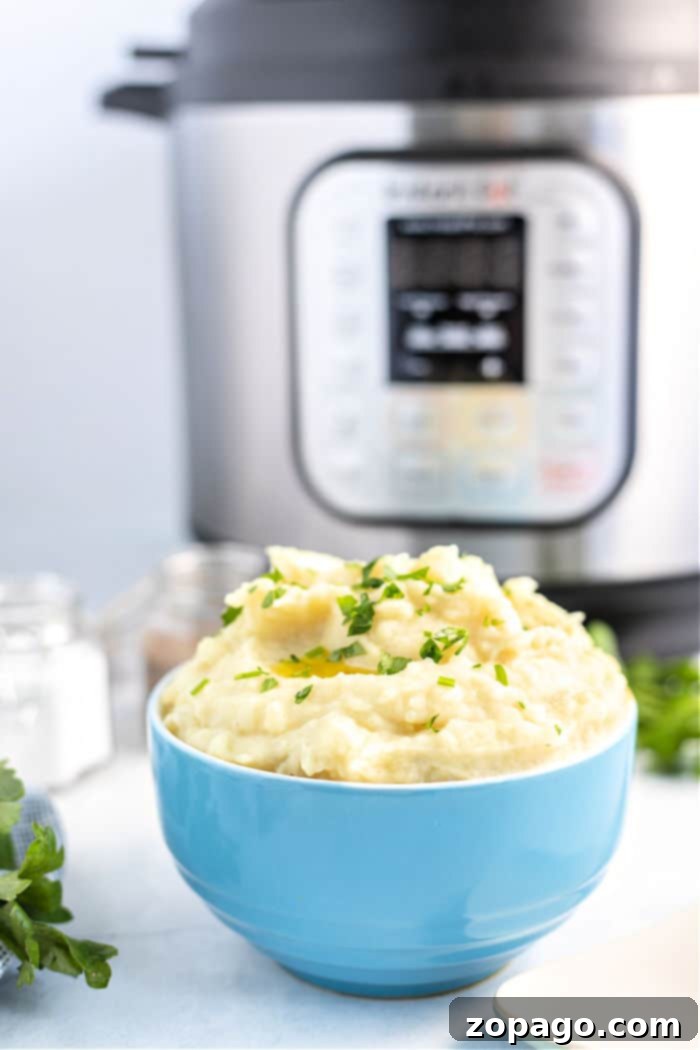 A bowl of creamy garlic mashed potatoes garnished with chopped herbs is in the foreground. In the background, there is an Instant Pot, its digital display glowing warmly on the kitchen counter.