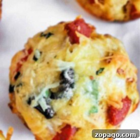 Close-up of a savory pizza muffin with visible pieces of cheese, olives, green herbs, and red ingredients, possibly pepperoni or tomatoes.
