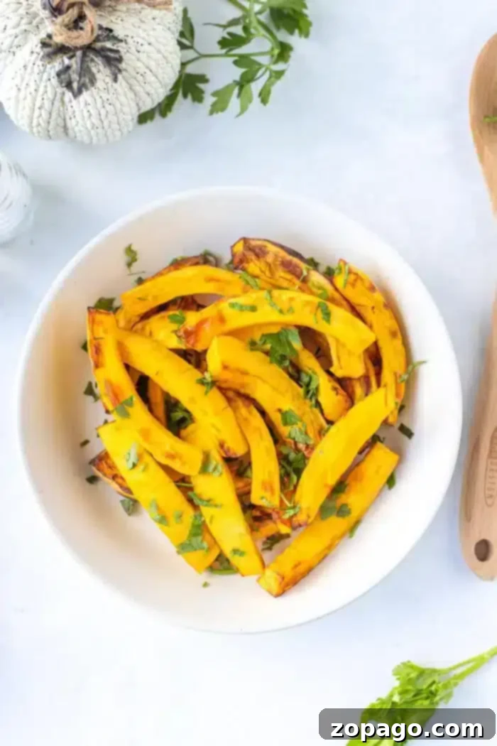 Crispy Air Fryer Pumpkin Fries 8 A white bowl contains roasted yellow squash garnished with chopped green herbs on a white surface. There is a wooden spoon on the right, and part of a decorative pumpkin and parsley can be seen in the top left corner, hinting at air fryer pumpkin slices to complete the autumnal scene.