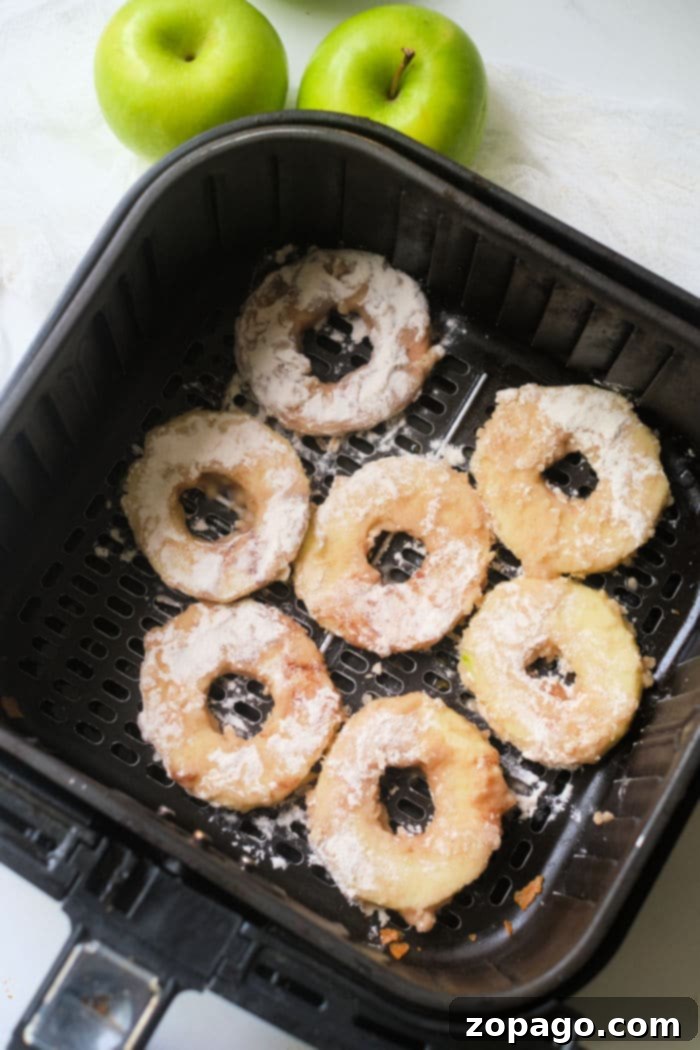 Perfect Air Fryer Apples 7 Crispy air fryer apple rings, perfectly golden and dusted with cinnamon sugar, are neatly arranged in the air fryer basket, with two fresh green apples nearby, symbolizing a healthy and delicious transformation.