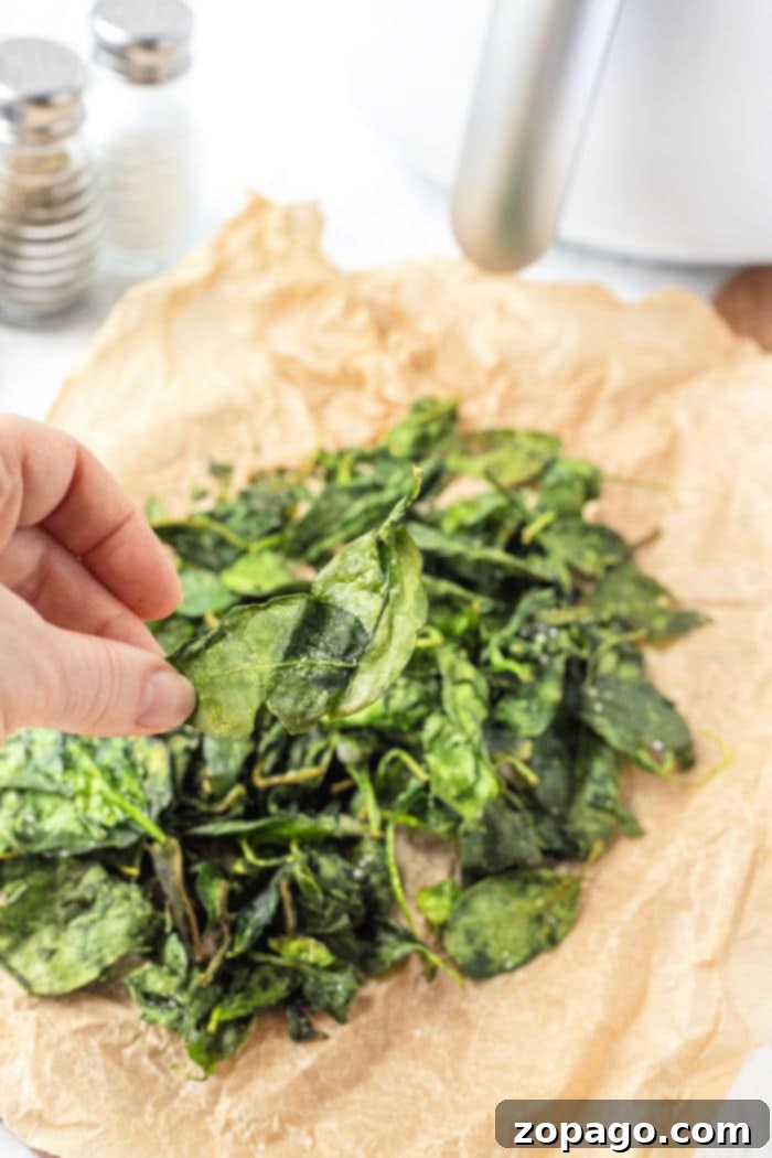 Crispy Air Fryer Spinach 10 A hand holds a crisp spinach leaf over a pile of perfectly air-fried spinach on parchment paper, showcasing the delicious result of the air fryer spinach method.