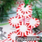 A Christmas ornament shaped like a gingerbread man is made from red and white peppermint candies, creating a whimsical, melting peppermint candy effect. It hangs by a green ribbon, with green pine branches in the background and holly designs on paper beneath it.