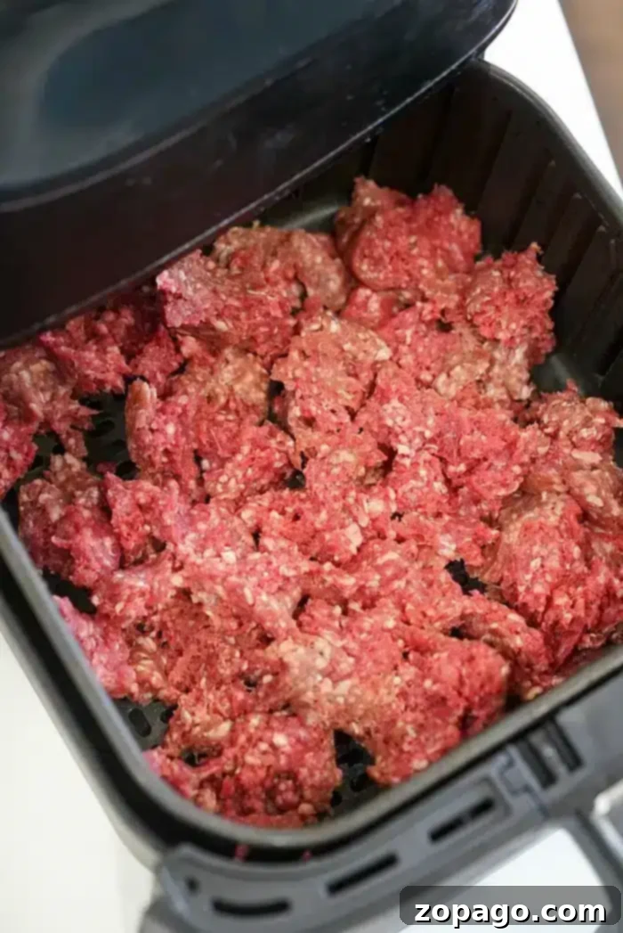 Perfect Air Fryer Beef 3 Ground beef is spread out in an air fryer basket, ready to be cooked. The air fryer lid is partially visible at the top of the image, perfect for those exploring new air fryer beef recipes.