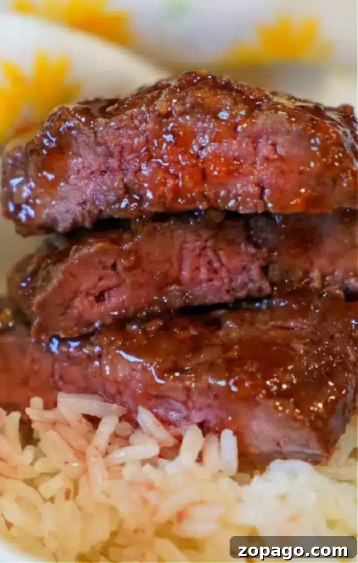Perfect Air Fryer Beef 5 A close-up shows three slices of barbecued beef with a shiny glaze stacked on top of each other, placed on a bed of white rice. The meat appears tender and moist with a slightly charred exterior, reminiscent of popular air fryer beef recipes. The background hints at a colorful, patterned tablecloth.