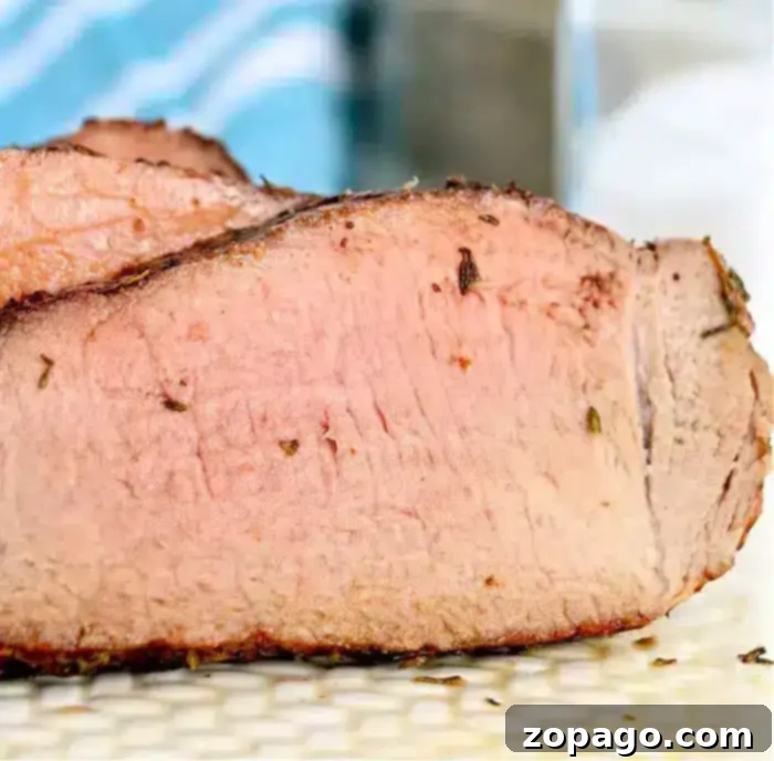 Perfect Air Fryer Beef 6 A close-up view of a thick, sliced piece of cooked meat with a pink interior and slightly charred outer edge, perfect for air fryer beef recipes. The background is slightly blurred, showing a blue and white striped cloth and a surface.
