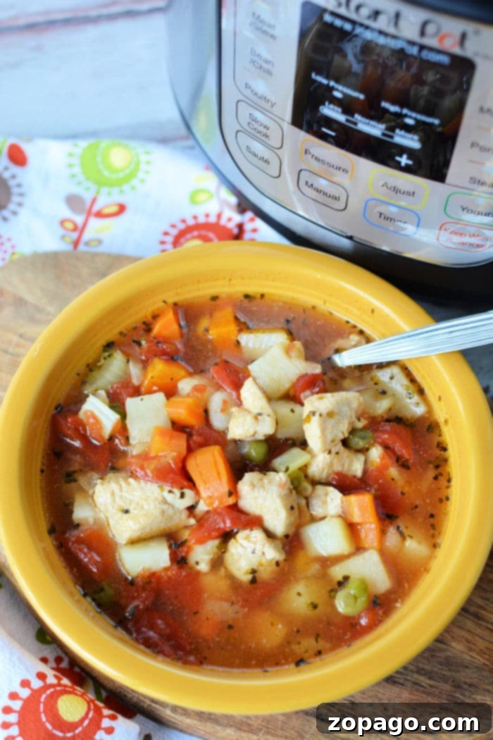 Hearty Instant Pot Chicken Vegetable Soup 7 A yellow bowl brimming with Instant Pot chicken vegetable soup features tender chicken, diced potatoes, carrots, peas, and tomatoes. A spoon rests in the bowl while an Instant Pot sits in the background on a floral patterned cloth.