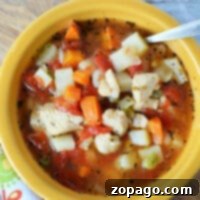 A yellow bowl of Instant Pot chicken vegetable soup brimming with diced potatoes, carrots, tomatoes, and peas, with a spoon resting inside. The bowl is placed on a decorative cloth.