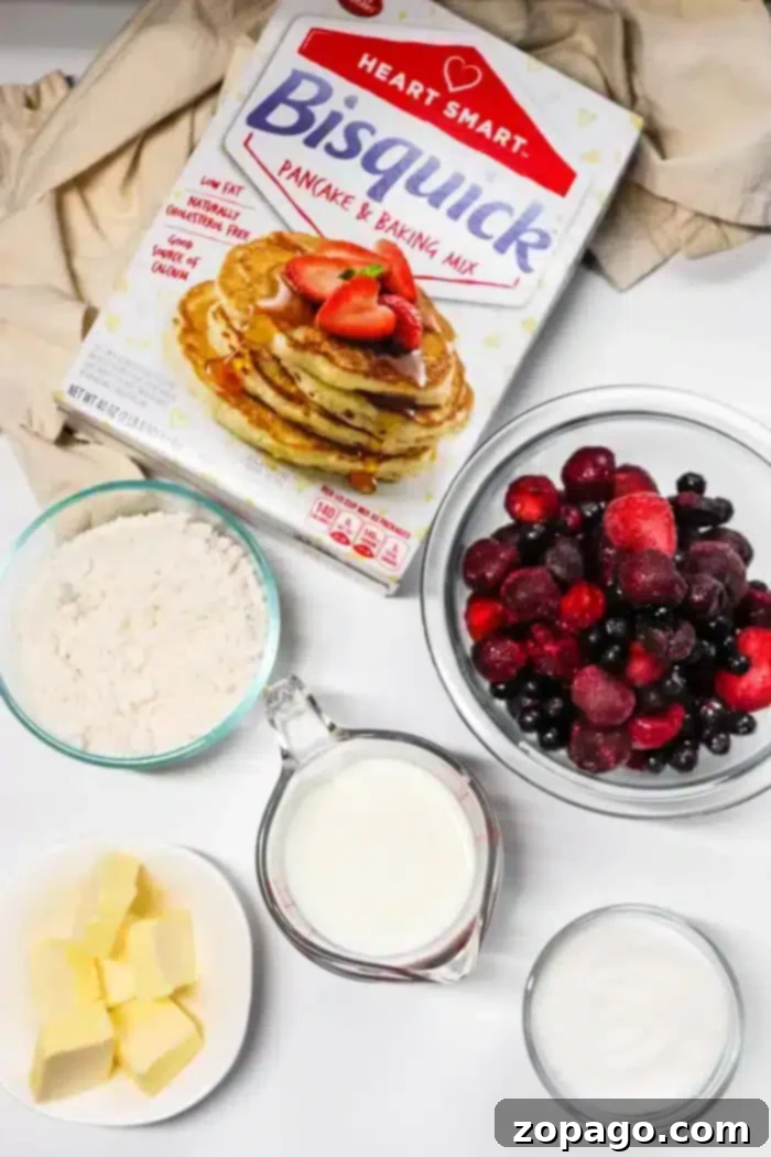 Easy 5 Ingredient Bisquick Blackberry Cobbler 3 A box of Bisquick pancake mix surrounded by bowls of flour, mixed berries for a blackberry cobbler, a cup of milk, a dish of butter, and a container of sugar, all arranged on a white surface with a beige cloth background.