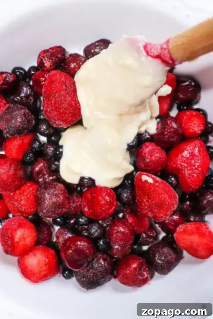 Easy 5 Ingredient Bisquick Blackberry Cobbler 5 A bowl of assorted berries, including strawberries, blueberries, and blackberries, topped with a dollop of creamy batter, resembling a Bisquick cobbler. A wooden spoon is partially dipped into the mixture.