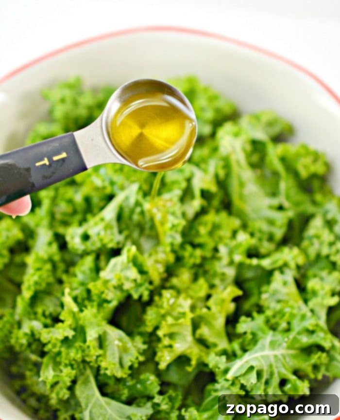Perfectly Crispy Oven Kale Chips 3 A tablespoon of olive oil is being drizzled over fresh kale in a white bowl, setting the stage for delicious baked kale chips.