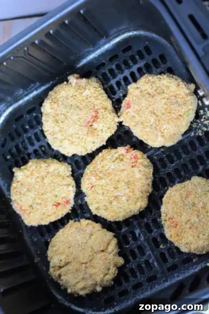 Golden Crispy Air Fryer Crab Cakes 6 Five perfectly breaded and round air fryer crab cakes are nestled within the perforated basket, poised for cooking. Each patty showcases a uniformly golden-brown crumb coating, promising a delicious crispiness. The dark, nonstick surface of the air fryer basket provides a striking contrast, highlighting the inviting appearance of the crab cakes.