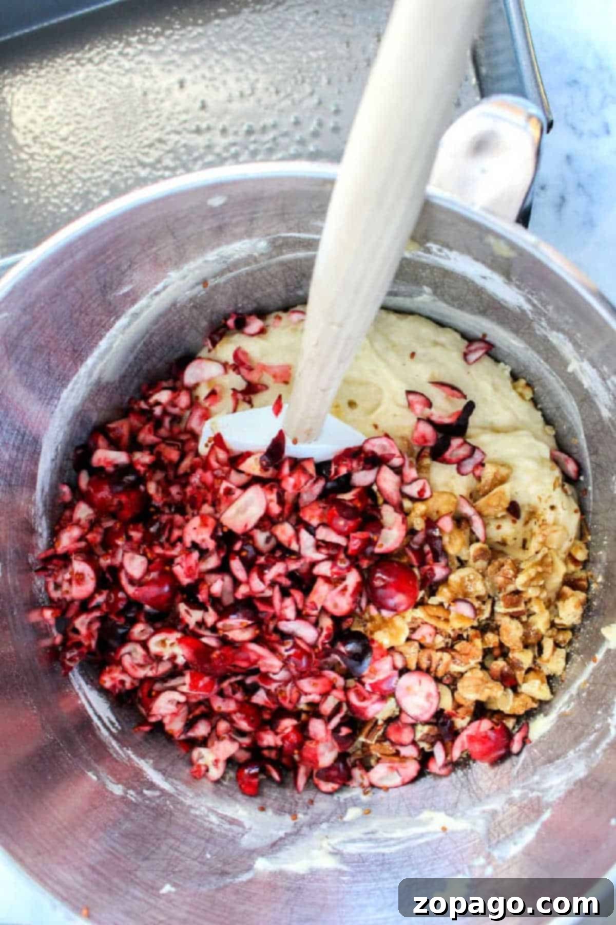 Golden Cranberry Banana Loaf 4 Freshly chopped cranberries and walnuts are being gently folded into a creamy banana bread batter in a shiny metal bowl using a spatula, preparing for a delightful bake.