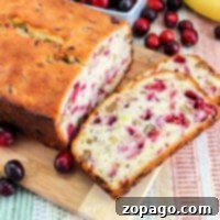 Sliced banana cranberry bread with walnuts rests on a wooden board, surrounded by fresh cranberries and a perfectly ripe banana, showcasing a delicious homemade treat.