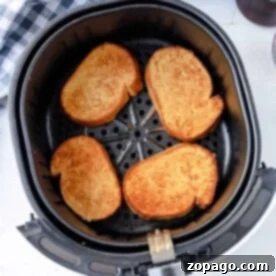Perfect Air Fryer French Toast 7 Four pieces of French toast sit perfectly toasted in an air fryer basket, with a checkered cloth nearby.