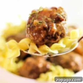 Slow Cooker Meatballs Smothered in Gravy 6 A fork with juicy crockpot meatballs and noodles, garnished with fresh herbs, stands out against a blurred background.