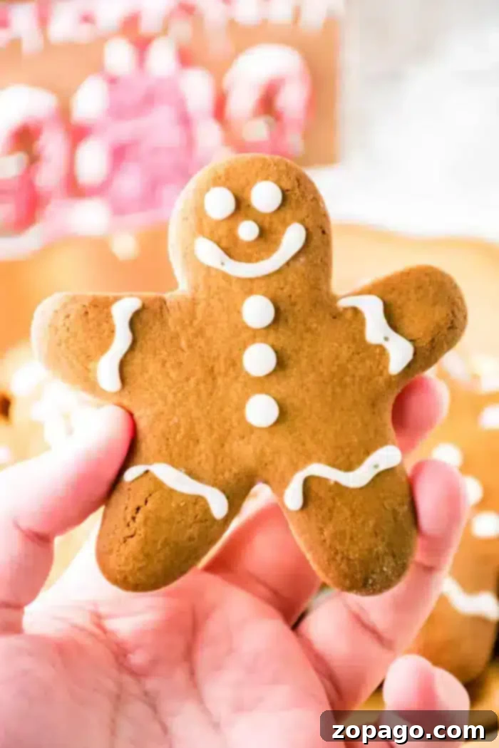 Perfectly Spiced Gingerbread Dough 2 A hand holds a gingerbread cookie, crafted from spiced gingerbread dough, shaped like a person and adorned with white icing for eyes, mouth, buttons, and outlines. Other similar cookies softly blur into the background.