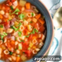 A bowl of healthy minestrone soup with pasta, vegetables, and herbs, simmered to perfection in a crockpot, sits next to a gold spoon on a patterned napkin.
