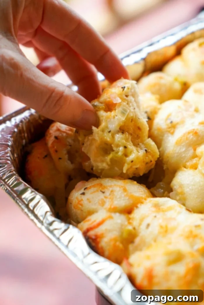 Smoked Traeger Appetizers 3 A hand picks up a piece of smoked, cheesy baked bread from a foil tray filled with golden, fluffy rolls. The bread appears slightly crispy with a rich, melted cheese topping.