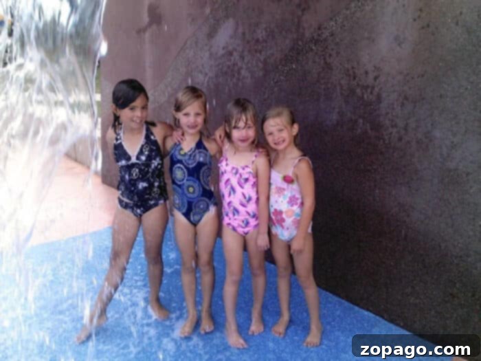 Splashing Good Times at Lake Skinner 3 Four young children in swimsuits stand and smile under a water feature at what appears to be the Lake Skinner splash pad. A water stream is visible on the left, with the kids standing on a blue surface against a textured wall.
