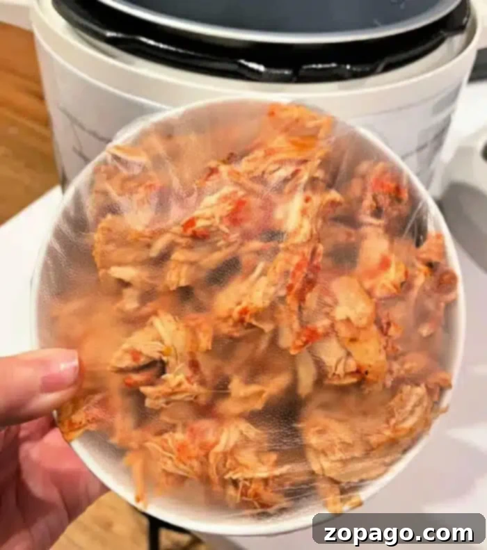 Instant Pot Leftover Chicken Makeovers 2 A person holds a plastic-covered bowl filled with seasoned, shredded chicken, perfect for delicious leftover chicken recipes. An Instant Pot can be seen in the background, ready to create delightful, quick one-pot dishes from these tender morsels.