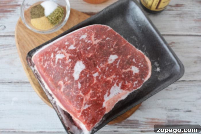 A block of frozen roast sits on a black tray, with a bowl of seasonings and Worcestershire sauce nearby.