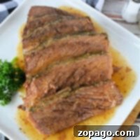 Perfectly Oven Cook a Frozen Roast 7 Sliced roast beef on a white rectangular plate, garnished with fresh parsley. A checkered black and white napkin and a salt shaker are placed nearby on a wooden surface. Juices from the frozen roast in the oven are visible on the plate.