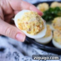 Hand holding a Crack Chicken Deviled Egg with a plate of more deviled eggs in the background.