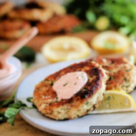 Air Fryer Tuna Patties 8 Two tuna patties rest on a plate, accompanied by a savory sauce and a lemon wedge, garnished with fresh herbs.