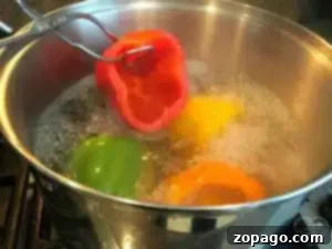 A stainless steel pot filled with boiling water on a stove holds green, yellow, and orange bell peppers. A metal utensil expertly demonstrates how to blanch bell peppers by submerging a red one into the water.