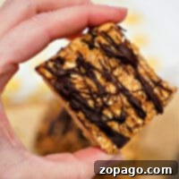 A hand holding a square oat bar drizzled with chocolate reveals a delicious blend reminiscent of chocolate peanut butter oatmeal bars.