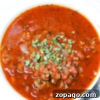 A bowl of Crockpot Venison Chili with minced meat, beans, and spices, garnished with chopped herbs, sits invitingly on a white plate accompanied by a spoon.