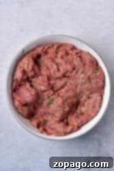 Hearty Venison Chili 5 Ground venison mixed with herbs in a white bowl on a gray surface.