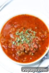 A bowl of Crockpot Venison Chili with minced meat, beans, and spices, garnished with chopped herbs, sits invitingly on a white plate accompanied by a spoon.