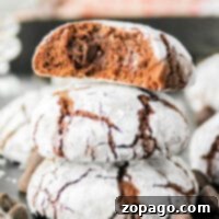 Three brownie mix crinkle cookies are stacked on a dark surface, with the top cookie broken in half to reveal the gooey chocolate inside. The cookies are dusted with powdered sugar, and chocolate chips are scattered around them.