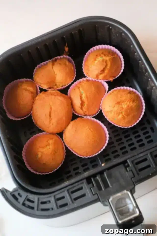 Easy Air Fryer Cupcakes 4 Seven golden-brown air fryer cupcakes baked in pink silicone cups, arranged neatly within the basket of a black air fryer, sitting on a light counter.