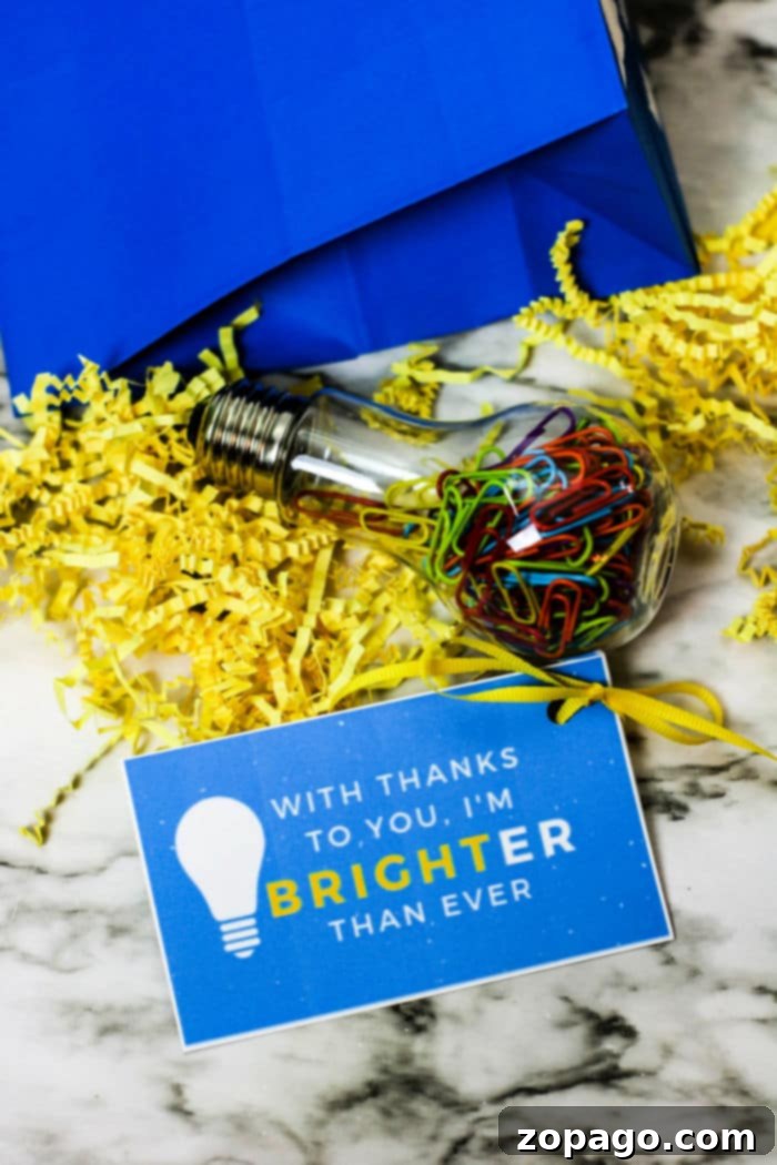 DIY Thank You Tags for Teachers 4 A blue gift bag on a marble surface contains yellow shredded paper and a clear bulb-shaped container filled with colorful paperclips. Alongside, a blue card with a lightbulb graphic reads, With thanks to you, I'm brighter than ever—perfect for attaching free printable teacher appreciation tags.