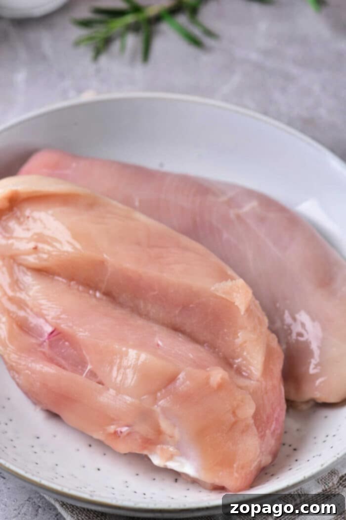 Zesty Italian Slow Cooker Chicken 3 A white plate containing two raw chicken breasts set on a gray surface with green herbs in the background. One chicken breast is slightly pinker in color, while the other appears less so. The chicken breasts have slight marbling and visible fat, ready to be transformed into Crockpot Chicken with Italian Dressing.