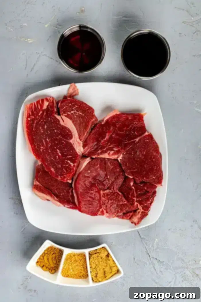 Signature Asian Steak Marinade 3 A square white plate with several pieces of raw red beef sits on a textured gray surface, suggesting a perfect base for an Asian steak marinade. Above the plate are two cups, one filled with a dark liquid and the other with a reddish liquid. Below the plate are three small white dishes with various spices.