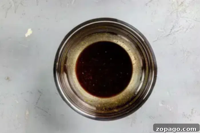 A stainless steel bowl containing a dark brown Asian Steak Marinade, resting on a textured grey surface, ready for use.
