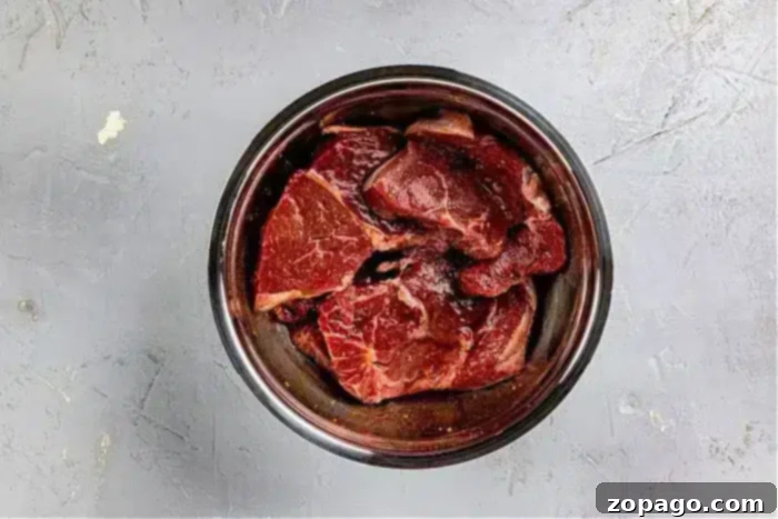 A metal bowl holds raw pieces of beef, perfectly marbled and fresh, ready to be immersed in an Asian steak marinade, positioned centrally on a gray textured surface.