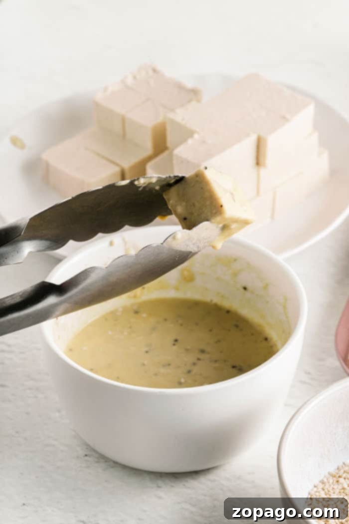 Crispy Tofu Bites 4 Tongs holding a tofu cube over a bowl of yellow sauce resemble the careful preparation of tofu nuggets, with more tofu blocks waiting on a plate in the background.