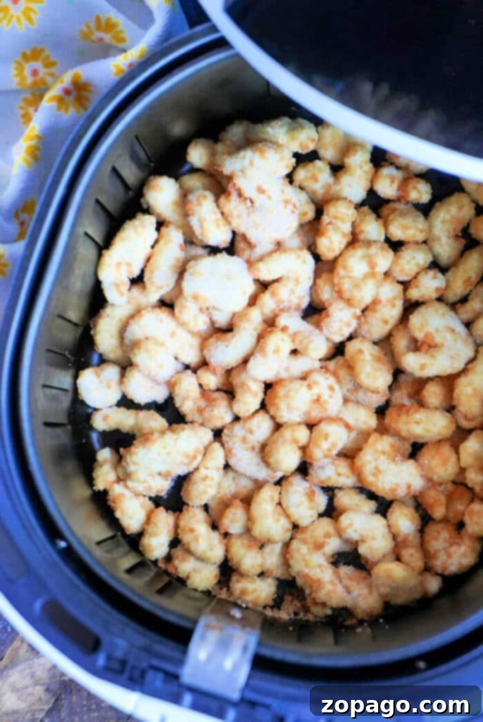 Perfectly Crispy Air Fryer Popcorn Shrimp 2 An air fryer basket brimming with crispy, golden-brown popcorn shrimp pieces sits enticingly on a surface, alongside a yellow floral-patterned cloth. It's the perfect setup to savor frozen popcorn shrimp in the air fryer next time.