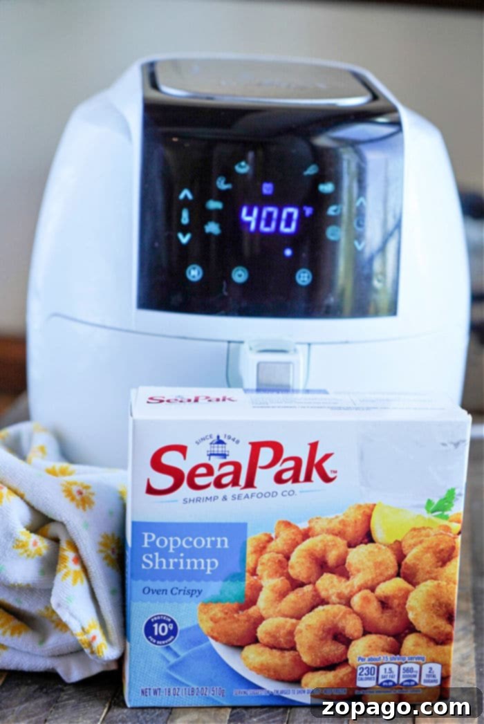 Perfectly Crispy Air Fryer Popcorn Shrimp 4 A white air fryer, ideal for cooking frozen popcorn shrimp, displays 400 on its digital panel. In front of it is a box of SeaPak Popcorn Shrimp, labeled Oven Crispy, with a lemon wedge pictured on the packaging. A yellow and white patterned cloth is nearby.